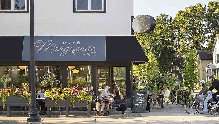 1 - Front exterior of Cafe Marguerite with people sitting at tables and two bicyclists arriving 1 - Front exterior of Cafe Marguerite with people sitting at tables and two bicyclists arriving